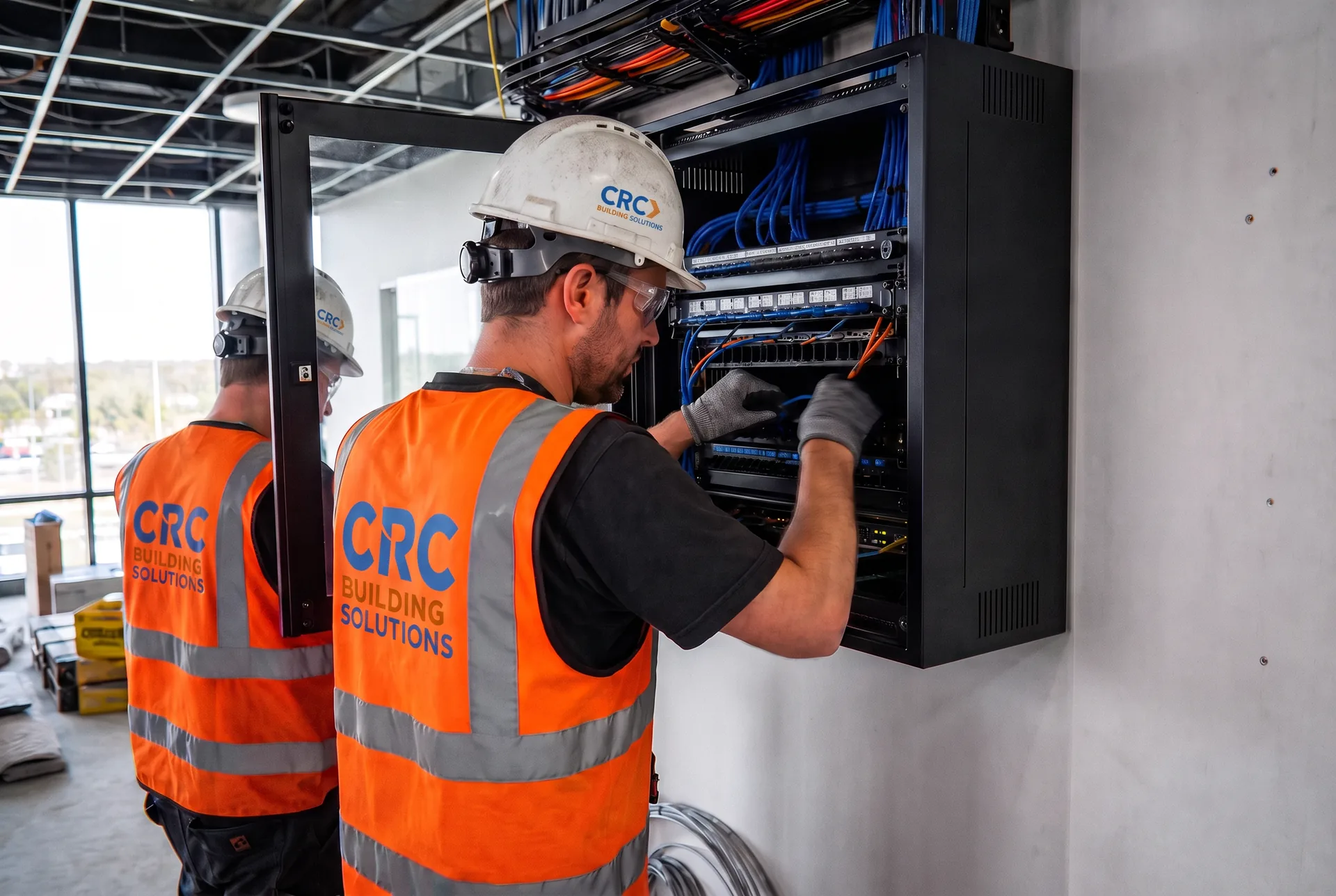 Two CRC Building Solutions low-voltage technicians in CRC-branded orange hi-vis vests and hard hats terminating blue Cat6 patch cabling inside a wall-mounted network rack during a commercial fit-out