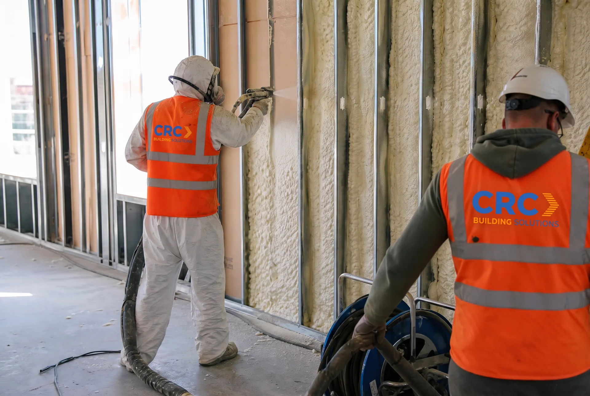 CRC Building Solutions insulation applicator in a hooded protective suit and CRC-branded hi-vis vest spraying closed-cell foam into steel-stud wall cavities while a second CRC crew member manages the hose reel